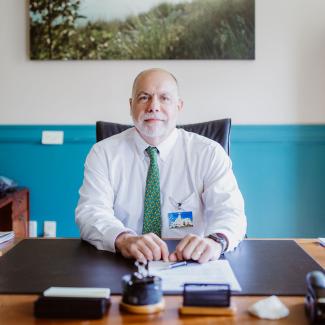 Rhode Island DEM Director Gray seated behind his desk looking to camera with a slight smile wearing white shirt and green tie. A photo of an estuary of Narragansett Bay is behind him on the way.